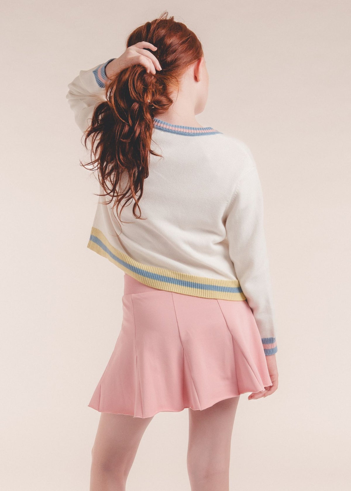 Tween girl from behind wearing a pink pleated skort and cream striped cardigan
