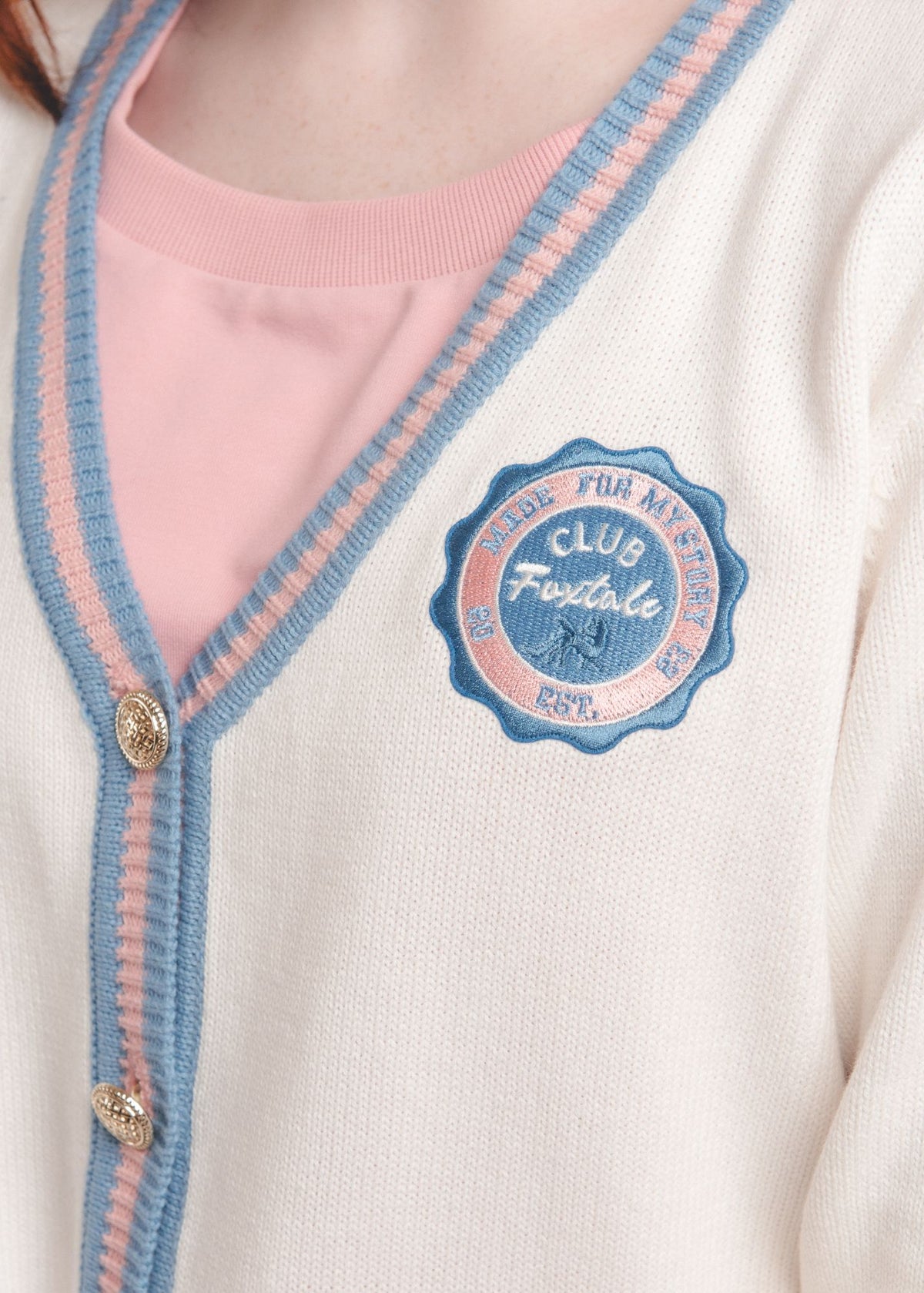Close-up of ivory cardigan with striped trim and an embroidered Club Foxtale patch