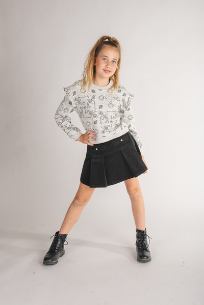 Tween girl wearing an ivory paisley bandana print sweatshirt with a ruffled shoulder and black pleated skirt