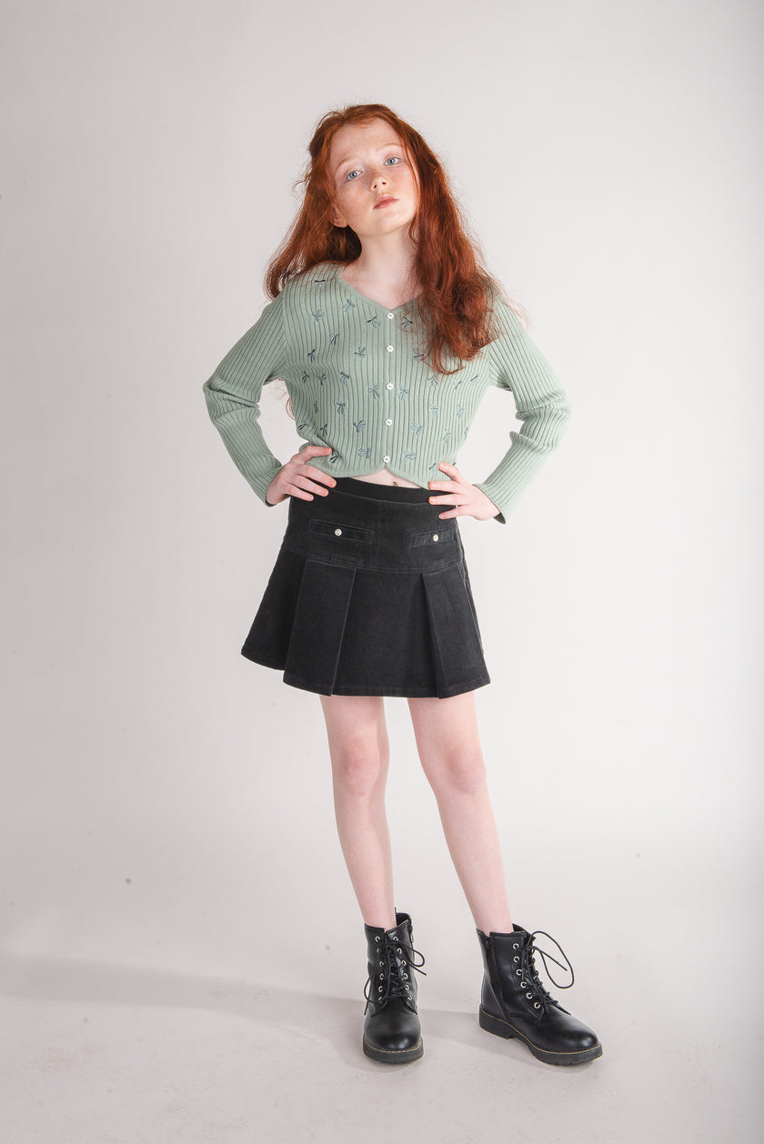 Tween girl wearing a black corduroy pleated skater skort with a sage green cardigan and boots