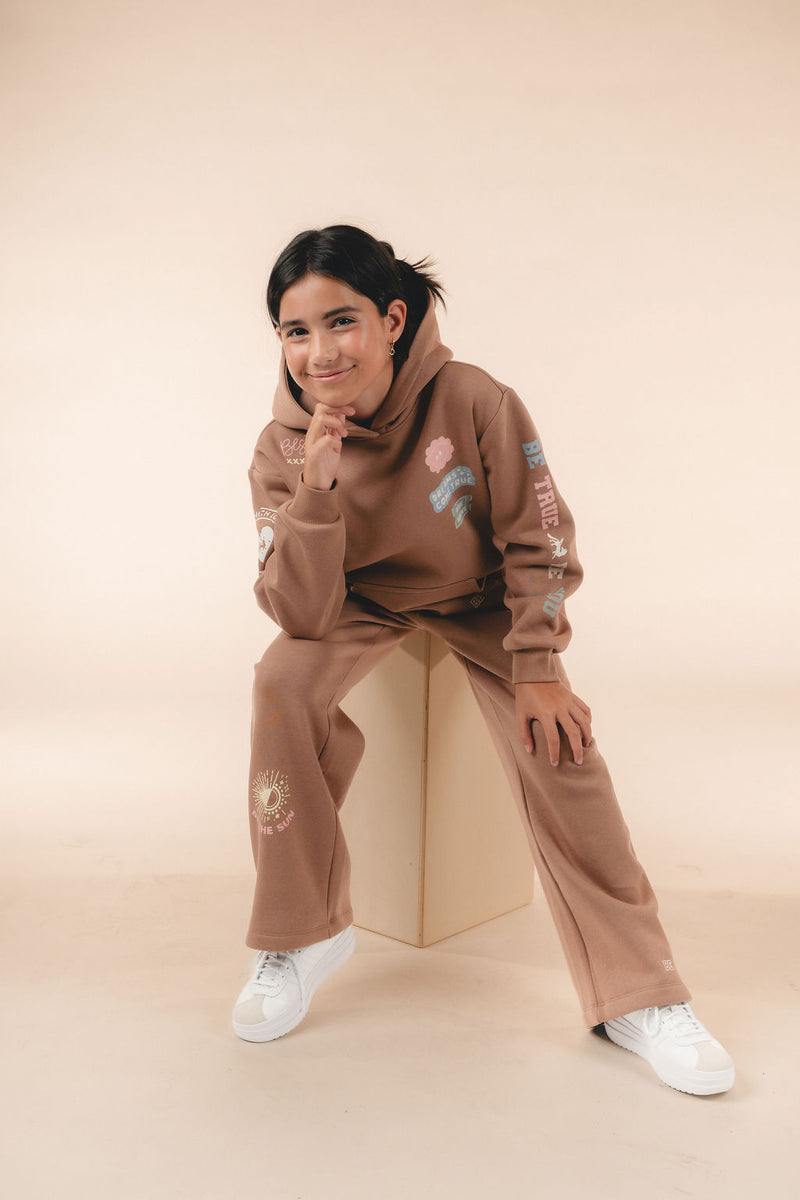 Girl in a brown fleece hoodie and wide-leg sweatpants with positive-message graphics, seated on a wooden box with her chin resting on her hand