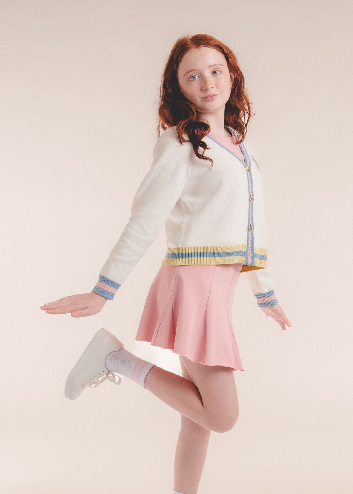 Tween girl jumping in a pink pleated skort with a cream striped cardigan