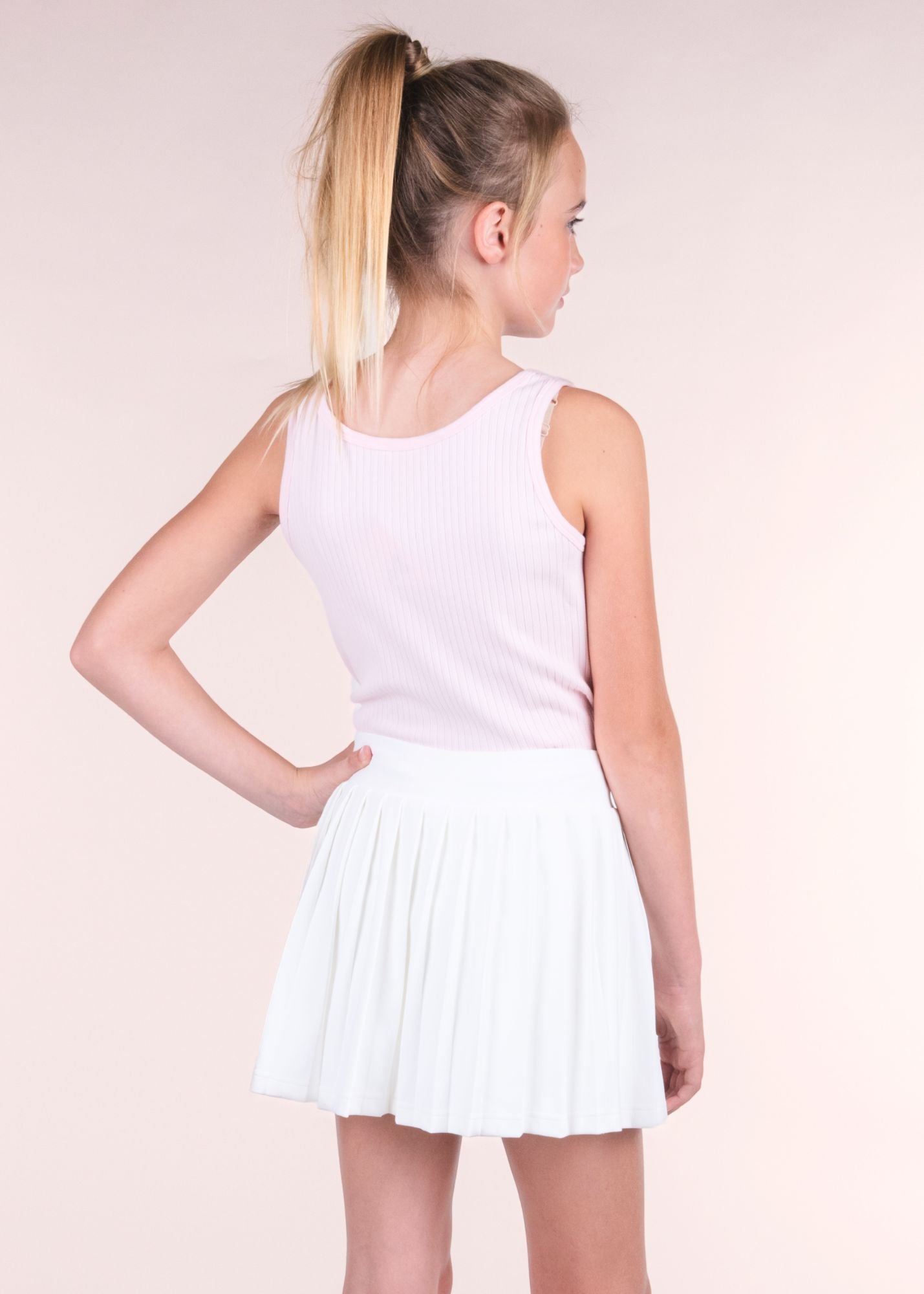 Tween girl wearing a blush pink ribbed tank top, back view, with white pleated skirt