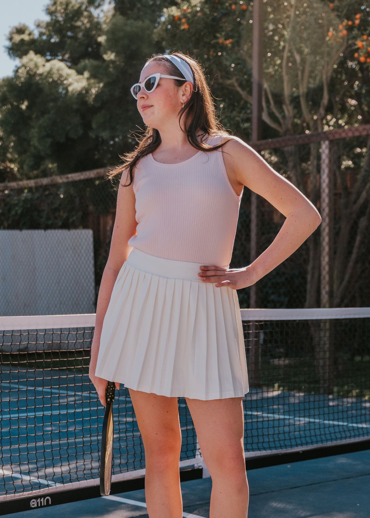 Tween girl wearing a blush pink ribbed tank top and white skirt, holding a tennis racket