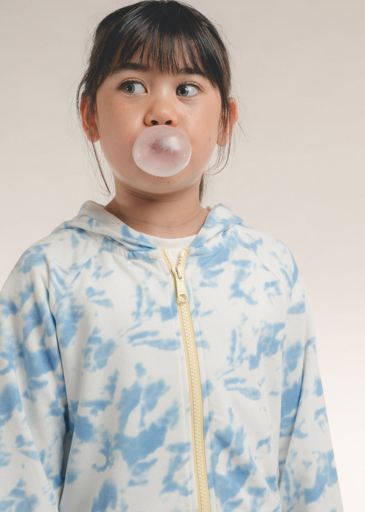 Tween girl blowing bubble gum in a blue-and-white tie-dye zip-up hoodie with hood down