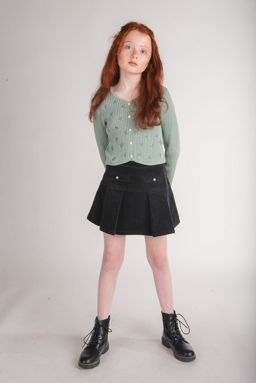 Tween girl wearing a sage green button-front sweater cardigan with black pleated skirt