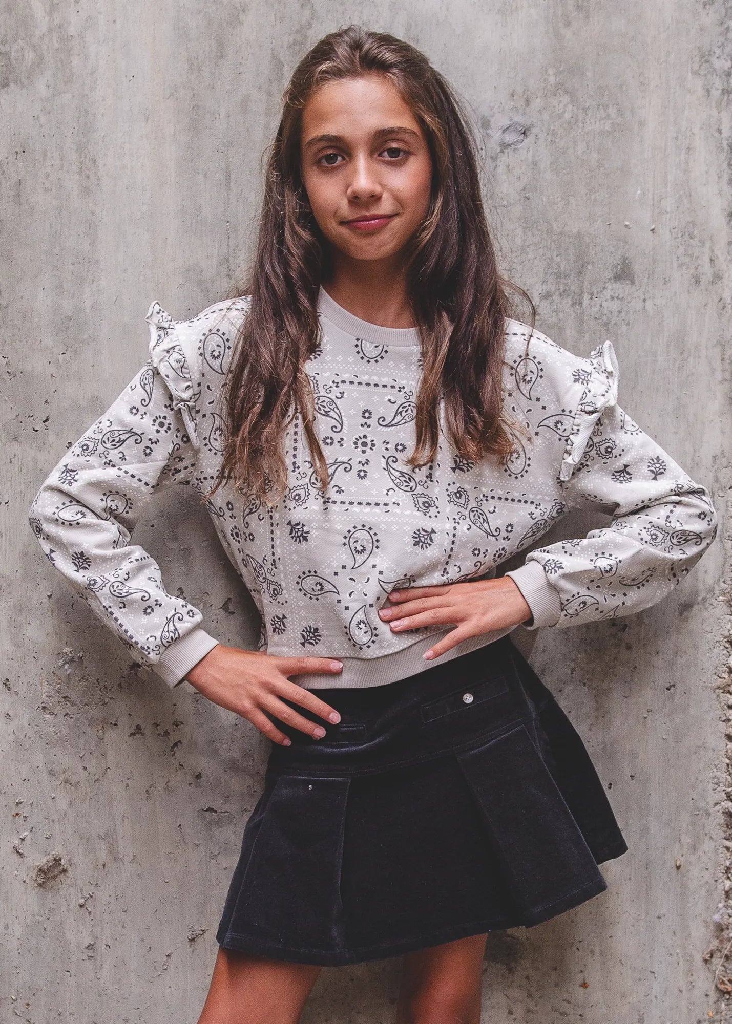 Young woman wearing a white blouse with black paisley patterns and a black skirt against a concrete wall.