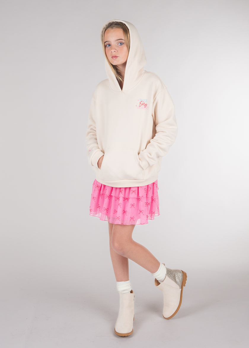 Tween girl wearing cream hoodie with hood up over a pink tulle bow print skirt
