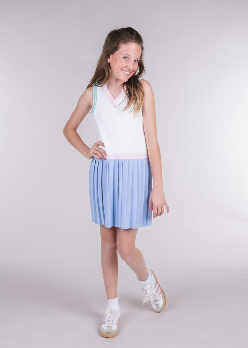 Tween girl in an ivory tennis dress with blue pleated skirt stands with one hand on hip